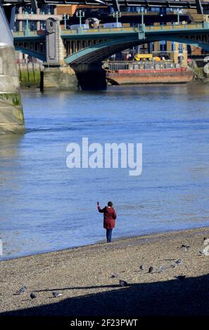 Londra, Inghilterra, Regno Unito. Donna Lone che prende un selfie sulla spiaggia del Tamigi a bassa marea Foto Stock