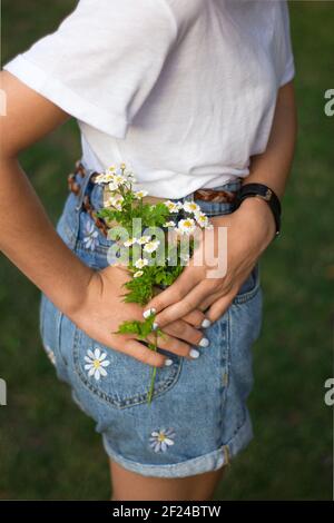 la ragazza adolescente irriconoscibile tiene un bouquet piccolo di margherite vicino alla tasca posteriore degli shorts del denim. Primo piano senza volto. Ciao estate. Giovani, tendono Foto Stock