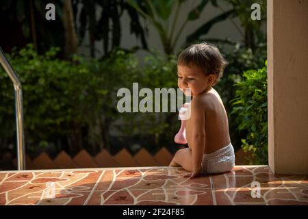 un affascinante bambino si siede sui gradini di una casa su sfondo verde giardino alla luce del sole. vista posteriore Foto Stock