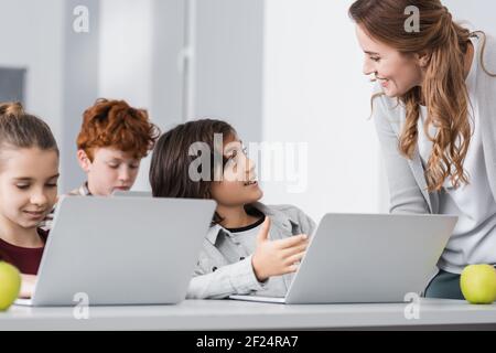 scolaro che indica il computer portatile vicino a un insegnante sorridente in classe Foto Stock