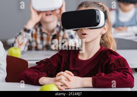 stupito gioco studentessa in vr visore vicino compagni di classe su sfocato sfondo Foto Stock