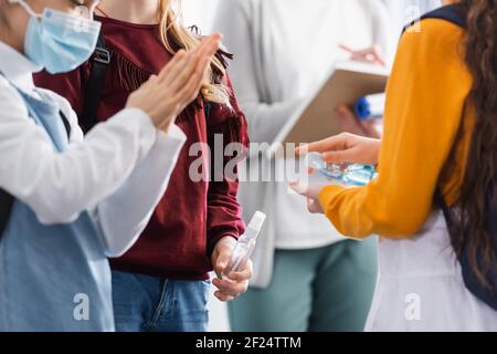 Studentessa che tiene la mano igienizzante vicino amico in maschera medica su primo piano sfocato Foto Stock