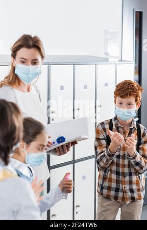 Ragazzo in maschera medica che tiene igienizzante per le mani vicino insegnante con termometro a infrarossi e notebook in corridoio Foto Stock