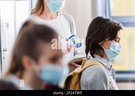 Insegnante che tiene il termometro a infrarossi e notebook vicino agli alunni in medicina maschere su primo piano sfocato Foto Stock