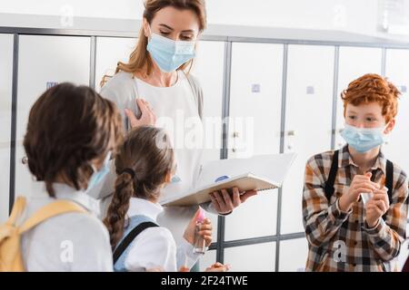 Alunni che tengono l'igienizzatore a mano vicino all'insegnante in maschera medica con notebook e termometro a infrarossi Foto Stock