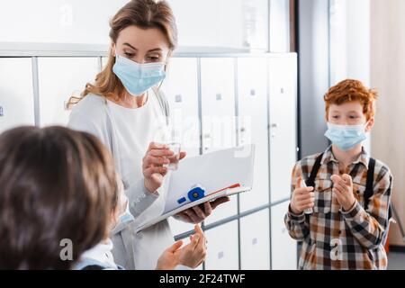 Insegnante con disinfettante, termometro a infrarossi e notebook in piedi vicino ai bambini in corridoio Foto Stock