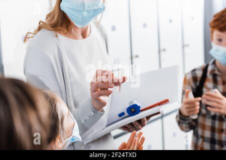 Insegnante con notebook e termometro a infrarossi che tiene vicino l'igienizzatore per le mani alunni in maschere mediche Foto Stock