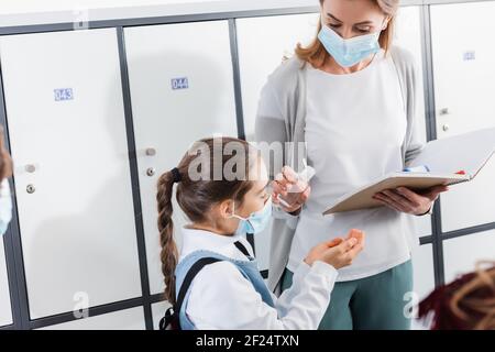 Insegnante con taccuino che tiene disinfettante vicino alla pupilla in maschera medica in sala Foto Stock