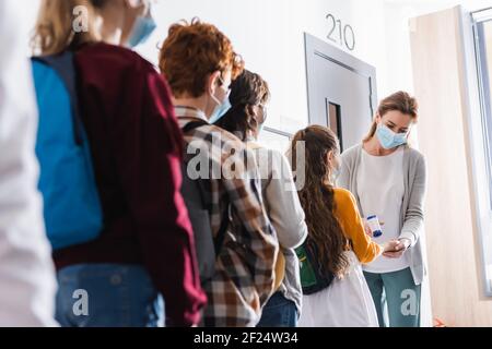 Insegnante in maschera medica utilizzando termometro senza contatto sulla pupilla vicino ai bambini in primo piano sfocato Foto Stock