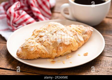 Croissant alle mandorle su un piatto servito con una tazza di caffè nero su un tavolo di legno. Vista in primo piano Foto Stock