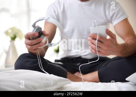 Uomo che tiene un cellulare e le cuffie bianche seduti sul letto con le gambe incrociate da vicino. Foto Stock
