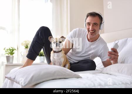 Uomo che ascolta la musica dal cellulare con le cuffie sorridenti il suo cane chihuahua in letto guardando e finestra in sfondo che entra il lig del mattino Foto Stock