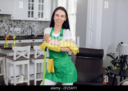 Giovane bella bruna sorridente caucasica donna, lavoratore di pulizia azienda in uniforme verde, posa sulla macchina fotografica con mop e panno durante la pulizia di Foto Stock
