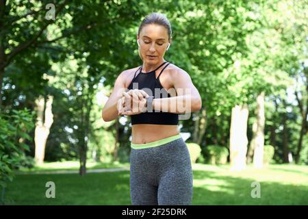 Jog al mattino. Bella donna sportiva in abbigliamento sportivo e auricolari guardando il suo orologio e controllando il risultato mentre jogging in un parco verde su un Foto Stock