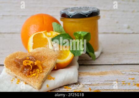 Toast a forma di cuore con marmellata d'arancia su un tovagliolo bianco con scorza di menta e arancio Foto Stock