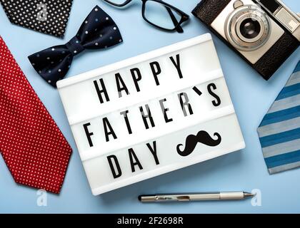 Happy Father's Day.Lightbox con la parola Happy Father's Day next a cravatte, cravatta di prua, fotocamera retrò, occhiali e penna su sfondo blu Foto Stock