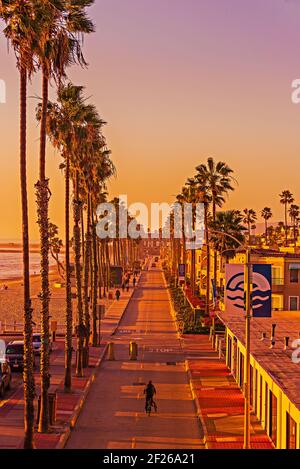 Strada cittadina che corre lungo la spiaggia fiancheggiata da palme e turisti al tramonto. Foto Stock