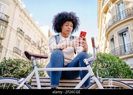 Donna Latina con acconciatura afro seduta su una panchina che guarda il suo cellulare. Foto di alta qualità Foto Stock