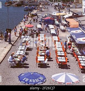 Portimão Algarve storico 1985 Vista aerea portoghese di archivio paesaggio urbano guardando in basso la gente del posto che cucina principalmente barbecue sardine pesce e serve cibo a gruppi di vacanze turistiche degli anni '1980 persone che mangiano all'aperto ai tavoli del caffè sul lungomare all'aperto sul soleggiato porto di pescatori di Portimão e banchina in Algarve Portogallo nell'immagine "The Way Were" dell'archivio anni '80 Foto Stock