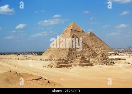 Grandi Piramidi di Giza si trovano su un cielo blu luminoso e sabbie del deserto giallo dorato, ma con la città invadente del Cairo sullo sfondo Foto Stock