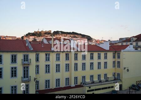 Vista del castello di Lisbona Sao Jorge dalla stazione ferroviaria di Rossio con edificio giallo in primo piano al tramonto, in Portogallo Foto Stock