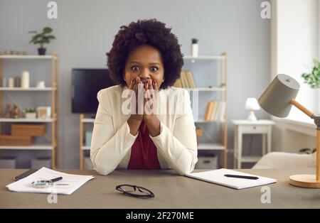 Ritratto di una donna afro-americana scioccata che ricopre la bocca con le mani siediti alla scrivania Foto Stock