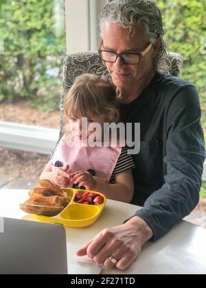 Nonno caucasico e sua nipote imparano a usare un computer portatile mentre si mangia il pranzo. Attività ricreative multigenerative per famiglie. Tempo di famiglia. Foto Stock