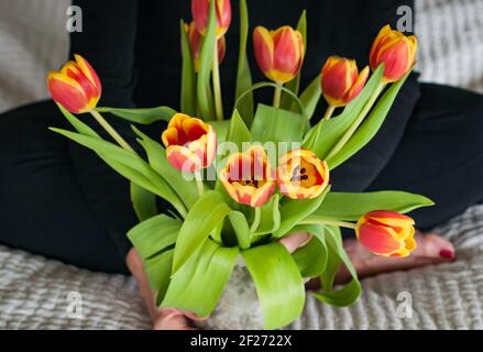 Vista ritagliata di una donna seduta in abiti neri che tiene un bouquet di tulipani arancioni e gialli. Mani che tengono i fiori in un vaso. Primo piano. Foto Stock