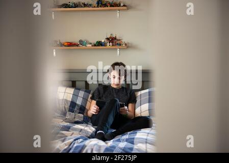 Guardando attraverso la porta il ragazzo Tween seduto sul suo letto con un tablet. Foto Stock