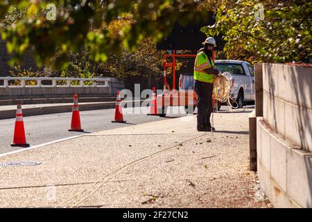 Washington DC, USA 11-02-2020: Un tecnico di elettricità che indossa giubbotto riflettente e casco sta installando il cavo sul lato della strada. Ci sono coni a des Foto Stock