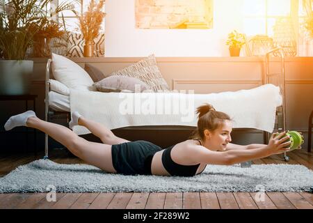 Una ragazza di fitness forte e bella sport in sportswear tiene la tavola con manubri sul pavimento a casa in la sua camera da letto luminosa e ariosa con un mi Foto Stock