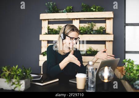 bella, giovane, bionda donna con eleganti e moderni occhiali neri si siede in un ufficio sostenibile e ha una riunione web online con una cuffia Foto Stock