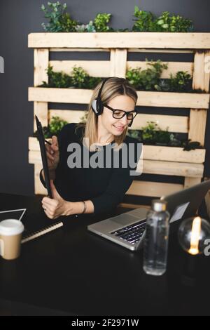 bella, giovane, bionda donna con eleganti e moderni occhiali neri si siede in un ufficio sostenibile e ha una riunione web online con una cuffia Foto Stock