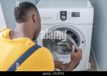 Uomo di servizio dalla pelle scura in camicia gialla che ripara la lavatrice Foto Stock