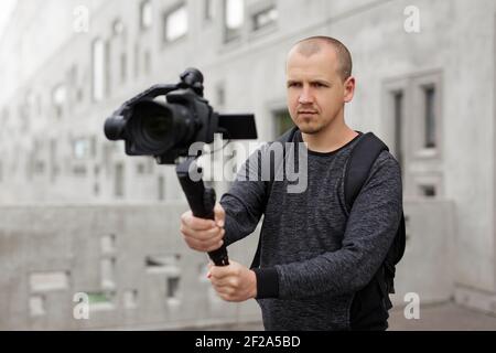 videografo professionale che scatta video utilizzando la moderna fotocamera dslr su 3 assi gimbal su sfondo grigio parete edificio in cemento Foto Stock