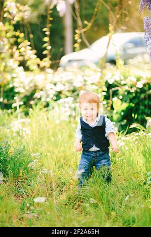 Il bambino carino dai capelli rossi sta in piedi su un'erba verde nel parco in una giornata di sole Foto Stock