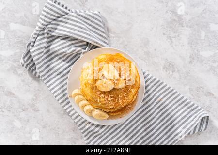 Pila di frittelle di banana al cocco appena sfornate guarnite con banane a fette, cocco tostato e sciroppo d'acero. Foto Stock