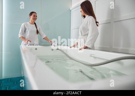 Donna allegra ed estetista in piedi vicino alla vasca da bagno nel salone del centro benessere Foto Stock