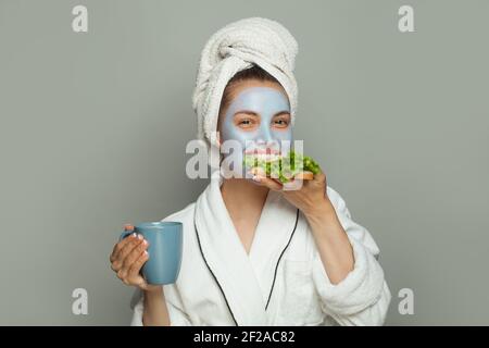 Bella donna in maschera viso cosmetica mangiare la colazione e bere caffè. Routine quotidiana femminile Foto Stock