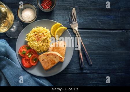 Gustoso corso di pesce con bistecche di tonno fritto, cavolo sabaudo, pomodori e limone serviti con vino, spezie e posate su tavola rustica di legno blu scuro, polpa Foto Stock