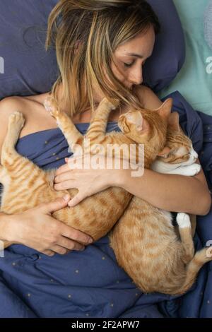 Giovane donna caucasica dai capelli biondi che giace sul letto della sua camera da letto, abbracciando i suoi due gattini tabbiani dai capelli arancioni. Foto Stock