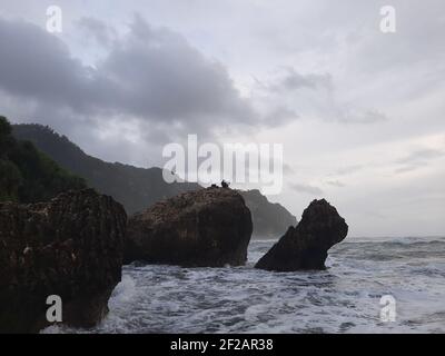 Un pescatore seduto su una formazione rocciosa mentre grandi onde lunghe fluiscono e fluiscono velocemente contro la scogliera e l'ampia spiaggia di sabbia a Parangtritis, Java. Foto Stock