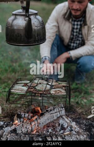 Il giovane friggete le verdure sulla griglia sul fuoco. Foto Stock