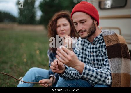 Accoppia in un plaid a scacchi arrostendo marshmallows sul fuoco vicino alla casa del rimorchio. Foto Stock