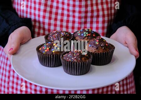 Ragazza che tiene un piatto di muffin. Nelle mani di un piatto da donna con dolci al cioccolato. Muffin con caramelle spruzzate. Giovane ragazza che cade in forno. Foto Stock