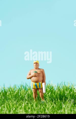 primo piano di un uomo in miniatura che indossa un costume da bagno a righe, tenendo un asciugamano in una mano e una tazza nell'altra mano, in piedi sull'erba, contro un blu Foto Stock