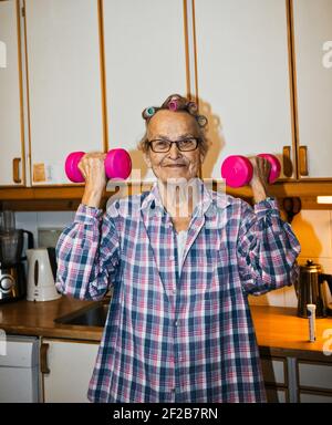 Allegra anziana anziana donna svedese che solleva i manubri rosa in cucina. Concetto di stile di vita, esercizi per anziani, stile di vita attivo Foto Stock