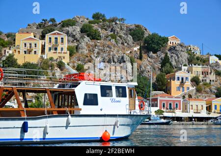 Paesaggio con case neoclassiche e una gita turistica in barca in primo piano nell'isola di Symi, Grecia Dodecanese. Foto Stock