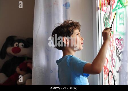 Un piccolo ragazzo serio tiene una spazzola nelle mani e disegna le immagini sulla finestra della sua stanza. Il bambino disegna un arcobaleno, una casa e un'astrazione. Foto Stock