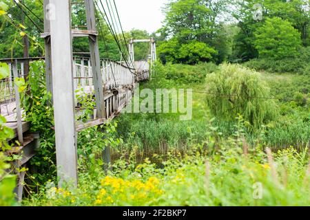 Vecchio ponte sospeso in ferro non funzionante su una vista laterale del fiume essiccato. Foto luminose e verdi succulenti. Un fiume surcresciuto. Foto Stock